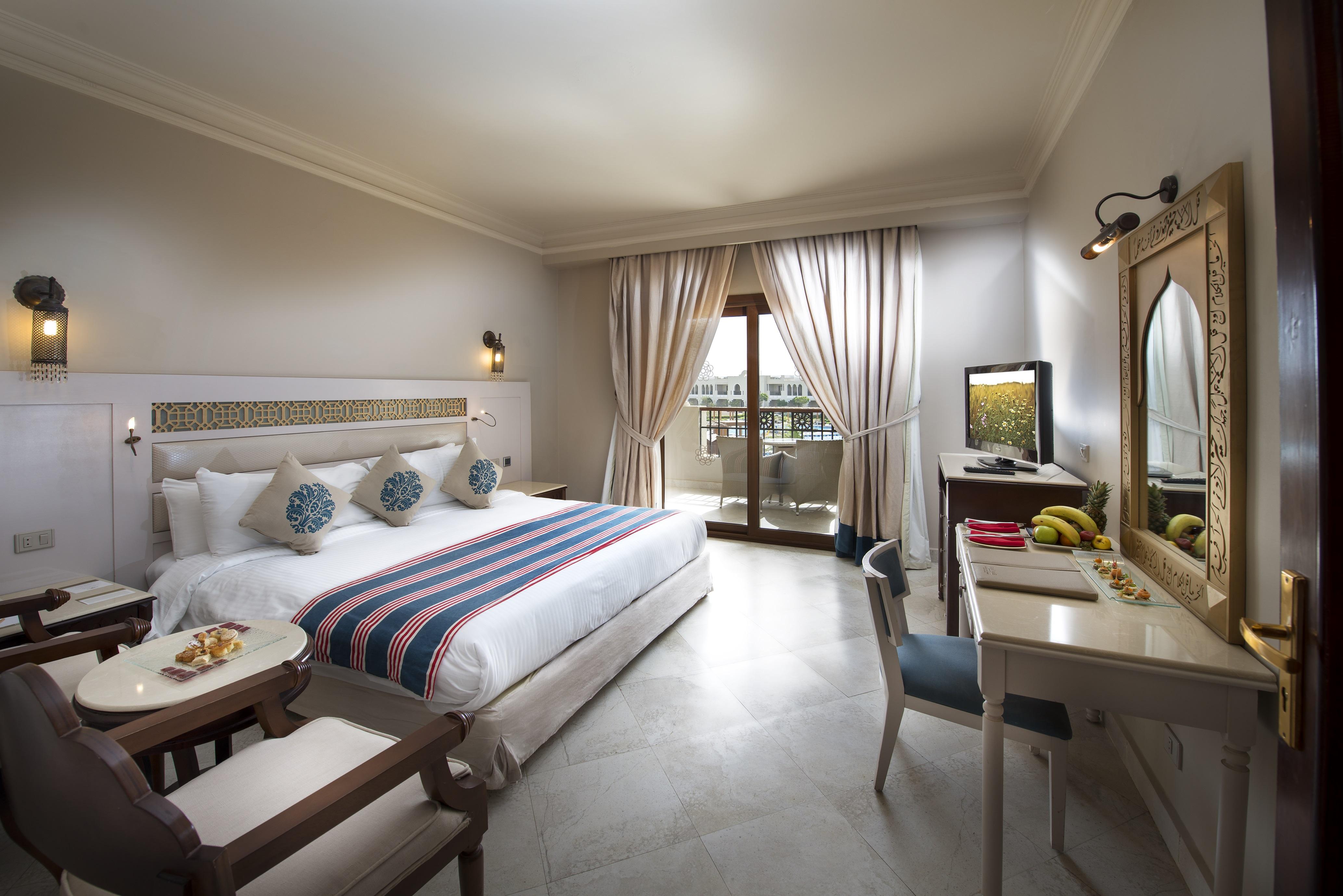 Sunrise Arabian Beach Resort Sharm el-Sheikh Exteriör bild The photo depicts a hotel room that appears stylish and well-furnished. It features a large bed with decorative pillows and a colorful blanket. There is a small seating area with armchairs and a round table, as well as a work desk with a chair. A tel