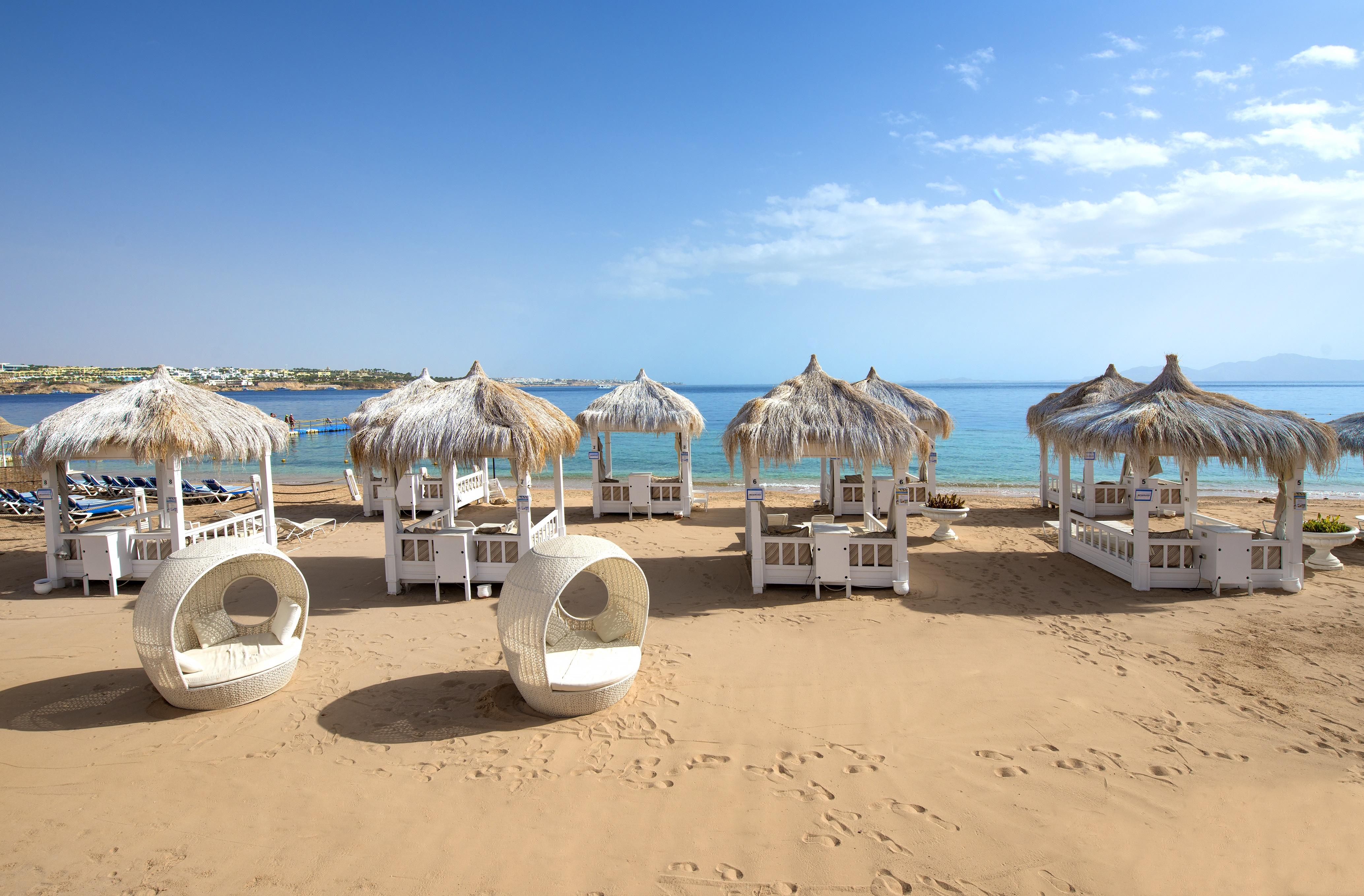 Sunrise Arabian Beach Resort Sharm el-Sheikh Exteriör bild The photo shows a picturesque beach scene featuring several stylish white cabanas with thatched roofs, set on golden sand. They are arranged neatly, with some circular lounge chairs nearby. In the background, the calm blue sea stretches out under a c