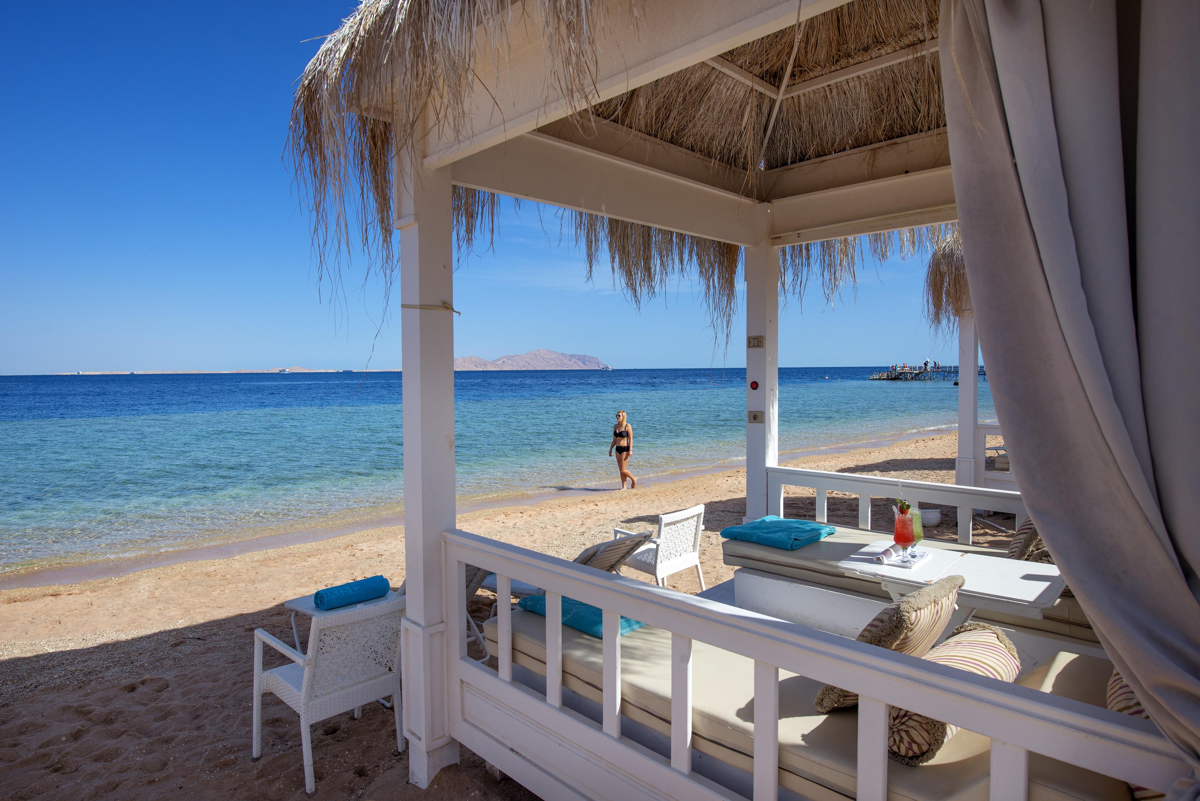 Sunrise Arabian Beach Resort Sharm el-Sheikh Exteriör bild The photo depicts a serene beach scene. In the foreground, there is a shaded seating area with white furniture and blue accents, creating a relaxing atmosphere. Behind the seating area, a sandy beach leads to calm, clear blue waters. A person is walk