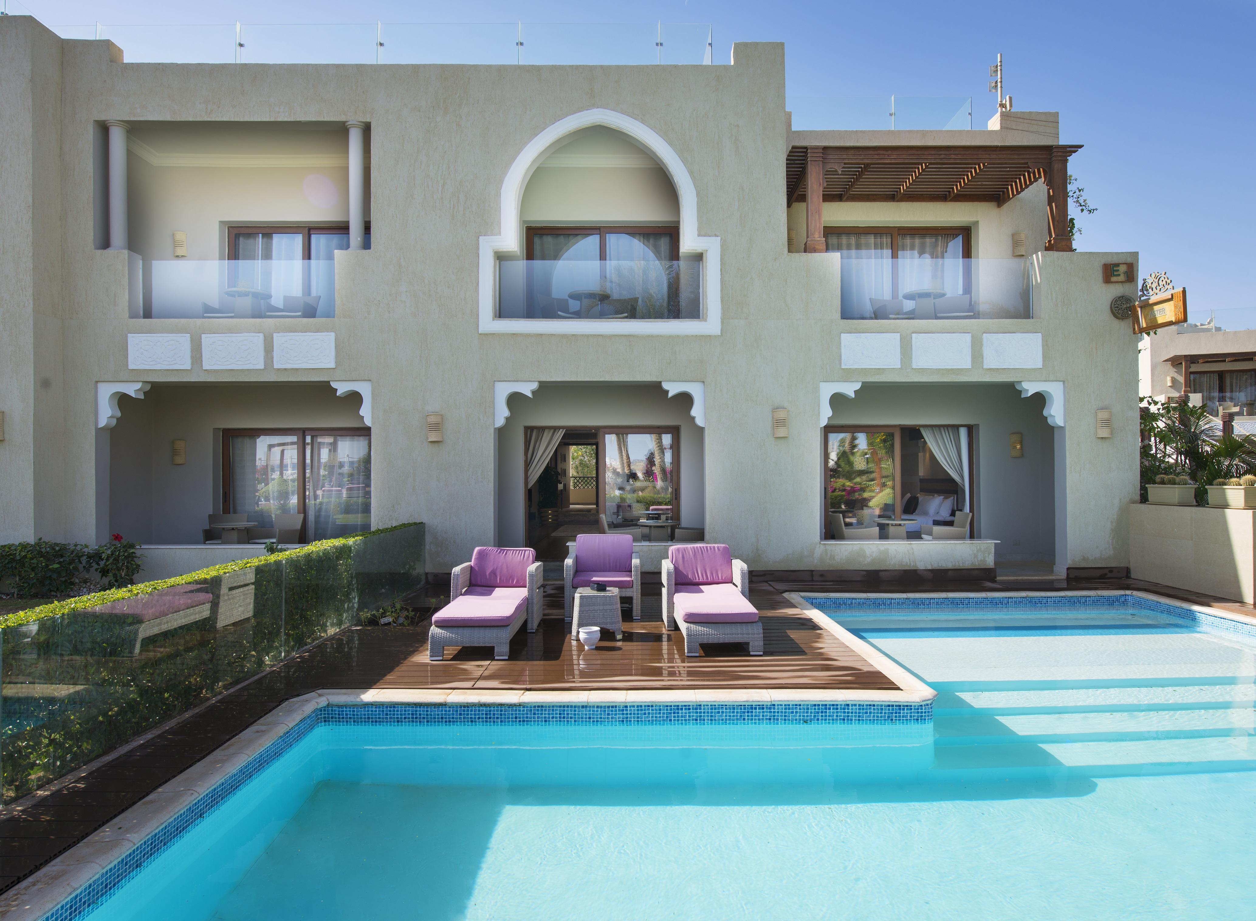 Sunrise Arabian Beach Resort Sharm el-Sheikh Exteriör bild The photo shows a luxurious modern villa with an elegant architectural style. In the foreground, there is a swimming pool that is clear and inviting, surrounded by a wooden deck. There are several lounge chairs with purple cushions positioned near th