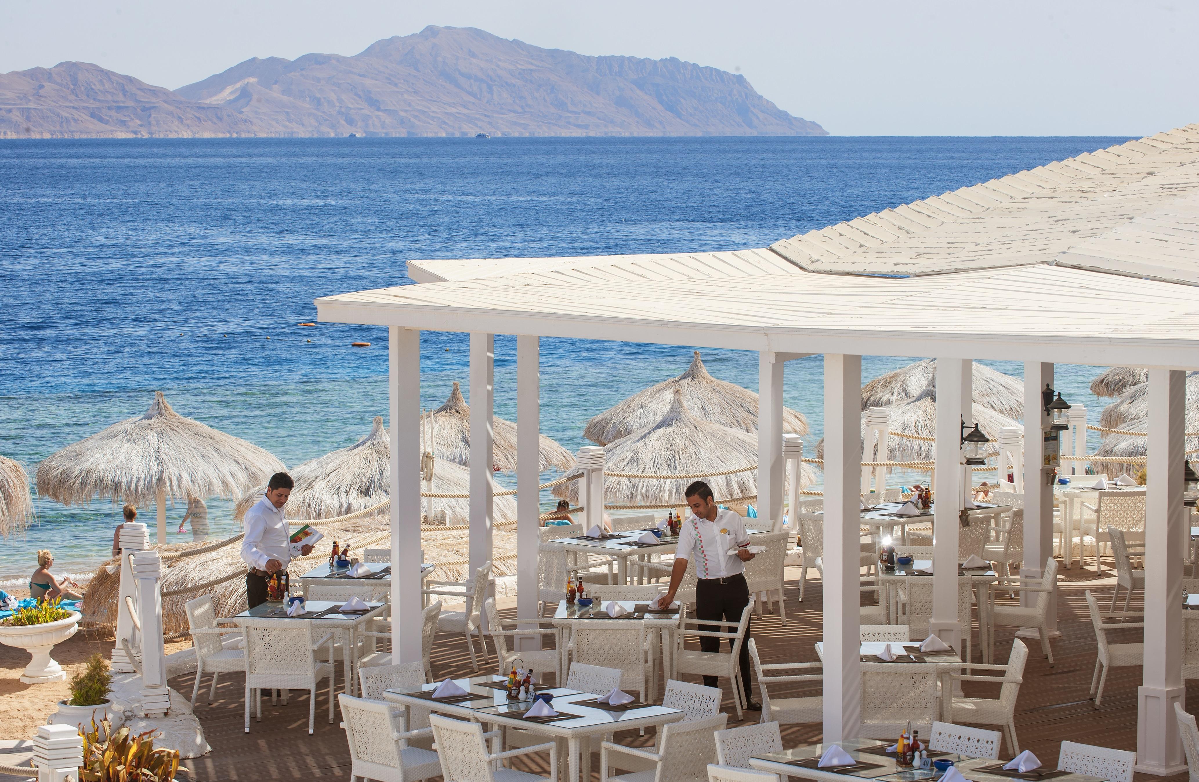 Sunrise Arabian Beach Resort Sharm el-Sheikh Exteriör bild The photo depicts a seaside restaurant with a beautiful view of the ocean and distant mountains. The outdoor dining area features white tables and chairs arranged under straw umbrellas for shade. Two waitstaff members are visible, attending to the ta