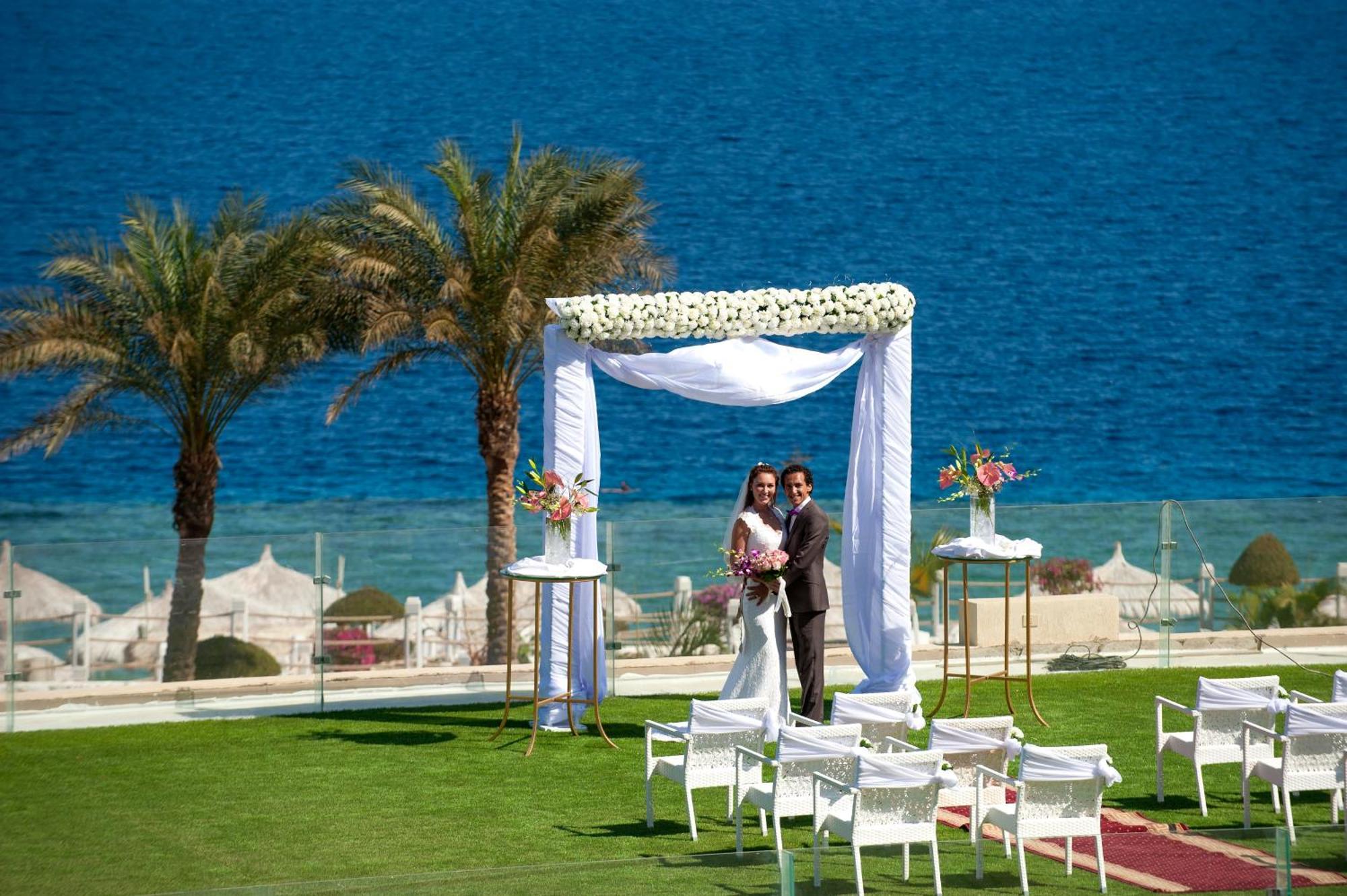 Sunrise Arabian Beach Resort Sharm el-Sheikh Exteriör bild The photo depicts a wedding scene set against a beautiful seaside backdrop. In the foreground, a couple is standing underneath a decorative wedding arch adorned with white fabric and flowers. The groom is in a dark suit, while the bride is dressed in