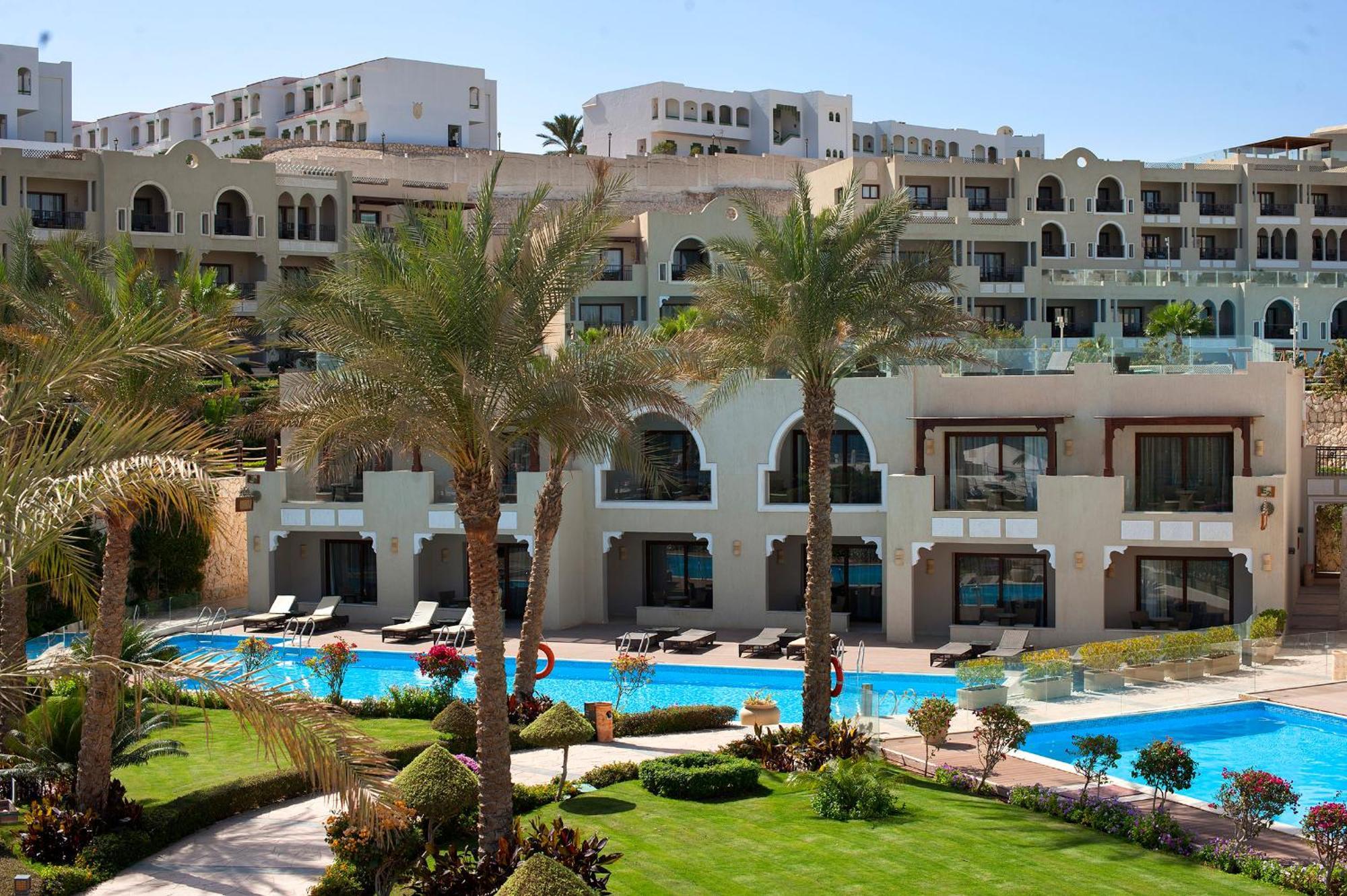 Sunrise Arabian Beach Resort Sharm el-Sheikh Exteriör bild The photo depicts a luxurious resort or hotel complex. In the foreground, there is a landscaped area with manicured lawns and palm trees. A swimming pool is visible, reflecting the clear blue sky. The background features several multi-story buildings